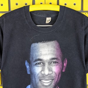 May include: Black t-shirt with a colorized image of a smiling man with short hair. The image is printed on the front of the shirt.