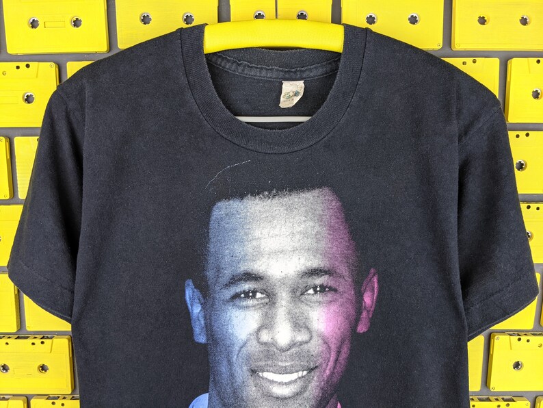 May include: Black t-shirt with a colorized image of a smiling man with short hair. The image is printed on the front of the shirt.