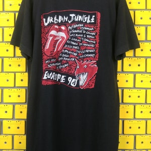 May include: Black t-shirt with a red and white graphic of a tongue and a dragon. The graphic includes the text "Urban Jungle" and a list of European cities and dates. The text "Europe 90" is also included.