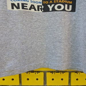Puede incluir: Camiseta gris con el texto "Coming Soon to a Stadium Near You" impreso en la parte delantera.