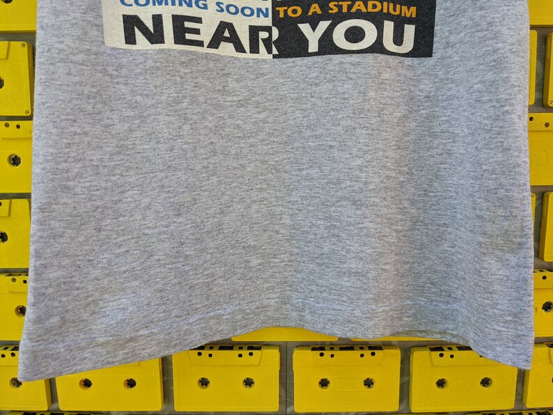 Puede incluir: Camiseta gris con el texto "Coming Soon to a Stadium Near You" impreso en la parte delantera.