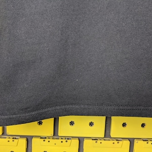 May include: Black t-shirt with a hemmed edge. The t-shirt is lying on a surface of yellow cassette tapes.