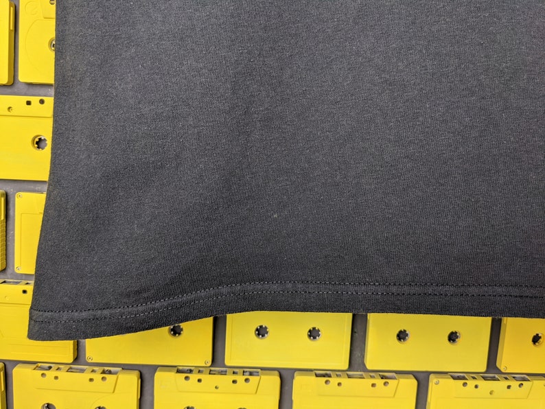 May include: Black t-shirt with a hemmed edge. The t-shirt is lying on a surface of yellow cassette tapes.