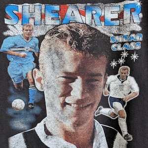 May include: Dark gray t-shirt featuring a portrait of a soccer player with the name "SHEARER" in blue and red. The text "SHEARER IS A GEORDIE" is printed at the bottom. Additional images of the player are also visible.