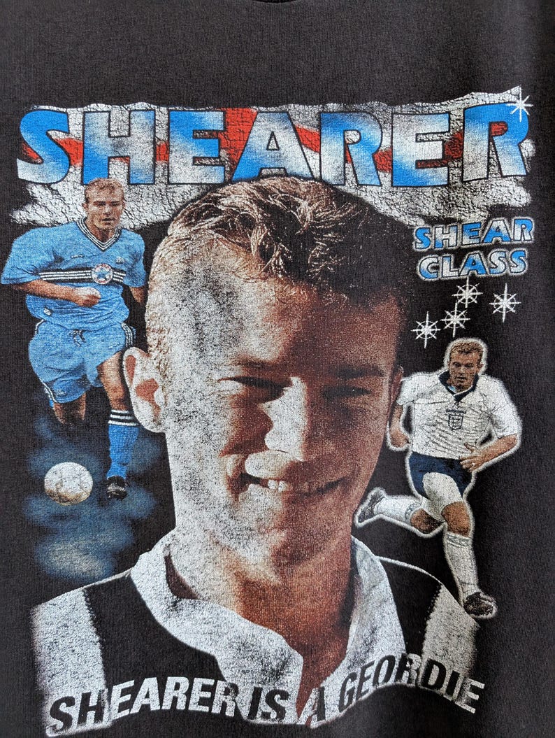 May include: Dark gray t-shirt featuring a portrait of a soccer player with the name "SHEARER" in blue and red. The text "SHEARER IS A GEORDIE" is printed at the bottom. Additional images of the player are also visible.