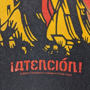May include: Black t-shirt featuring a yellow and red graphic of stick figures. The text "&iexcl;ATENCI&Oacute;N!" is printed in red below the graphic. The text "&copy; 2000 At The Drive In. Licensed to Cinder Block" is printed in white below the red text.