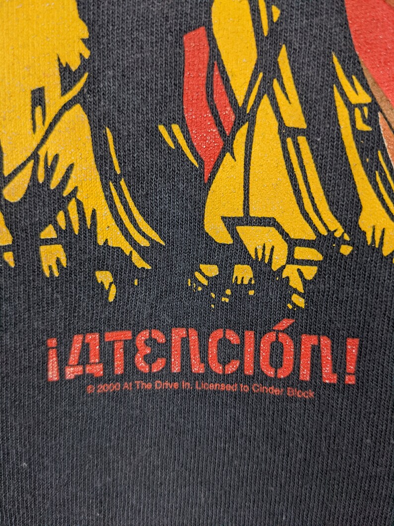 May include: Black t-shirt featuring a yellow and red graphic of stick figures. The text "&iexcl;ATENCI&Oacute;N!" is printed in red below the graphic. The text "&copy; 2000 At The Drive In. Licensed to Cinder Block" is printed in white below the red text.