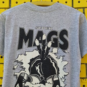 Puede incluir: Una camiseta gris con un estampado gr&aacute;fico en blanco y negro de un personaje de dibujos animados con el texto "SUPER MAGS" sobre el personaje.