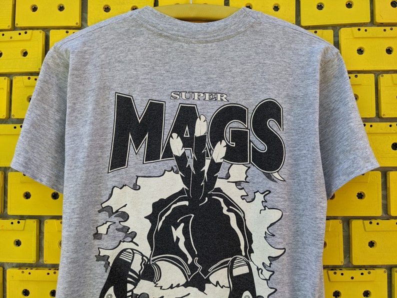 Puede incluir: Una camiseta gris con un estampado gr&aacute;fico en blanco y negro de un personaje de dibujos animados con el texto "SUPER MAGS" sobre el personaje.
