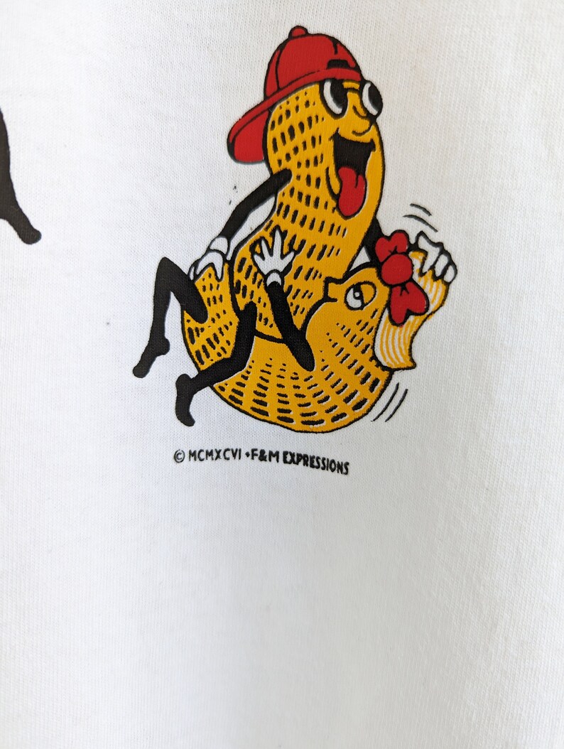 May include: A white t-shirt with a cartoon image of a peanut wearing a red baseball cap and sunglasses riding another peanut. The text "MCMXCVI - F&M EXPRESSIONS" is printed below the image.