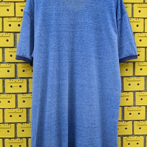 Puede incluir: Una camiseta azul de manga corta con un aspecto descolorido y texturado. La camiseta tiene un ribete azul marino alrededor del cuello y las mangas.