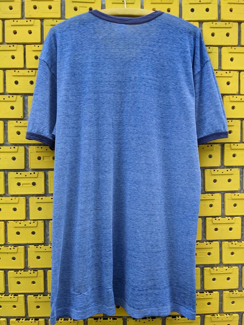 Puede incluir: Una camiseta azul de manga corta con un aspecto descolorido y texturado. La camiseta tiene un ribete azul marino alrededor del cuello y las mangas.