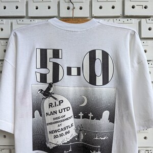 Pu&ograve; includere: Maglietta bianca con una stampa grafica in bianco e nero di una lapide con il testo "R.I.P. Man Utd Died of Embarrassment at Newcastle 20.10.96" e il numero "5-0" sopra la lapide.