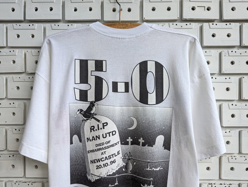 Pu&ograve; includere: Maglietta bianca con una stampa grafica in bianco e nero di una lapide con il testo "R.I.P. Man Utd Died of Embarrassment at Newcastle 20.10.96" e il numero "5-0" sopra la lapide.