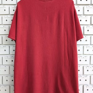 May include: A solid red t-shirt hangs on a wooden hanger. The short-sleeved shirt is made of a soft material and is displayed against a white wall decorated with cassette tapes.