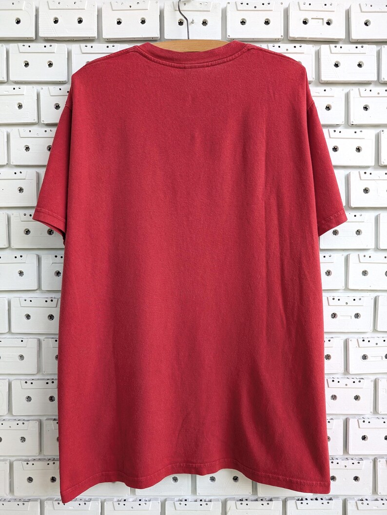 May include: A solid red t-shirt hangs on a wooden hanger. The short-sleeved shirt is made of a soft material and is displayed against a white wall decorated with cassette tapes.