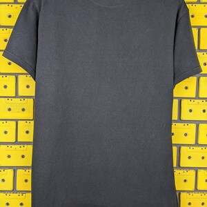 May include: A black t-shirt with a crew neck and short sleeves. The shirt is photographed against a background of yellow cassette tapes.