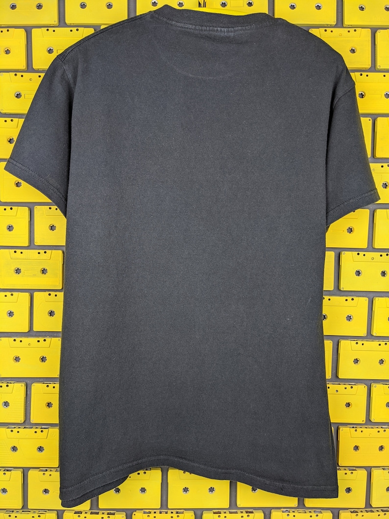 May include: A black t-shirt with a crew neck and short sleeves. The shirt is photographed against a background of yellow cassette tapes.