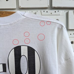 Pu&ograve; includere: Maglietta bianca a maniche lunghe con un design a strisce bianche e nere. Il design presenta un grande numero "0" con un rettangolo pi&ugrave; piccolo a sinistra. La maglietta ha diverse piccole macchie chiare.