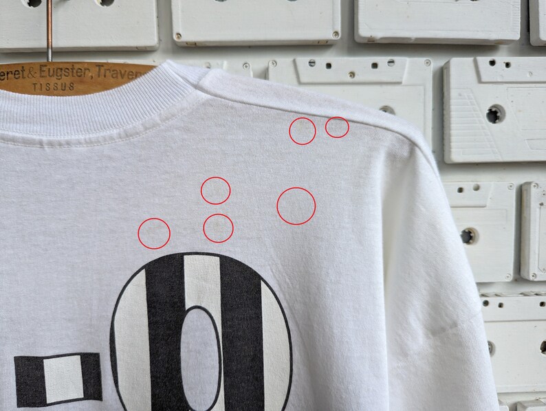 Pu&ograve; includere: Maglietta bianca a maniche lunghe con un design a strisce bianche e nere. Il design presenta un grande numero "0" con un rettangolo pi&ugrave; piccolo a sinistra. La maglietta ha diverse piccole macchie chiare.