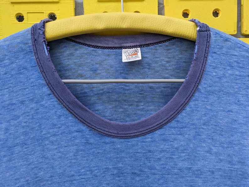 Puede incluir: Una camiseta azul con un cuello de canal&eacute; azul oscuro y una etiqueta de Hanes. La camiseta est&aacute; hecha de una tela suave y ligera.