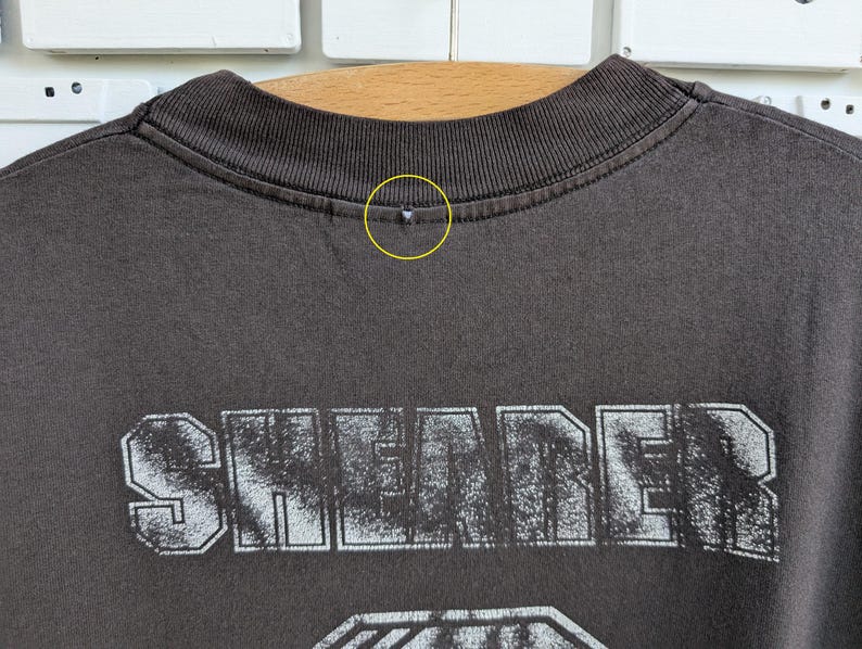 May include: Close-up of a dark gray t-shirt with the word "SHEARER" printed in a distressed, white font. The shirt's collar is ribbed and dark gray. The shirt appears to be made of a soft material.