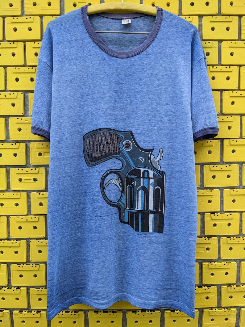 Puede incluir: Una camiseta azul con un gr&aacute;fico de un rev&oacute;lver en la parte delantera. La camiseta tiene un cuello redondo acanalado azul oscuro.