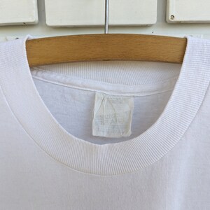 May include: White t-shirt with a ribbed neckline. The shirt is hanging on a wooden hanger.