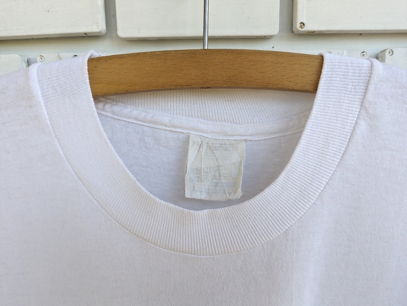 May include: White t-shirt with a ribbed neckline. The shirt is hanging on a wooden hanger.