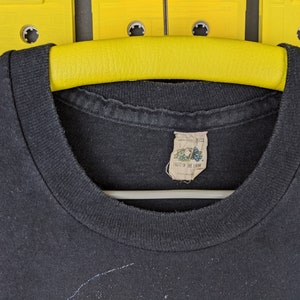 May include: Black t-shirt with a Fruit of the Loom tag. The t-shirt is hanging on a yellow hanger.