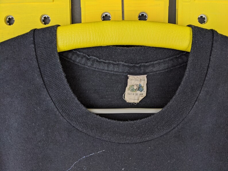 May include: Black t-shirt with a Fruit of the Loom tag. The t-shirt is hanging on a yellow hanger.