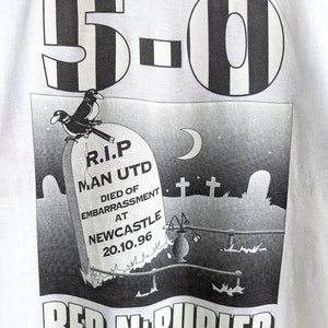Pu&ograve; includere: Una maglietta bianca con una stampa grafica in bianco e nero. La grafica raffigura una lapide con il testo "R.I.P. Man Utd Died of Embarrassment at Newcastle 20.10.96" e due uccelli appollaiati in cima. Il testo "5-0 Red N' Buried" &egrave; stampato sotto la grafica.