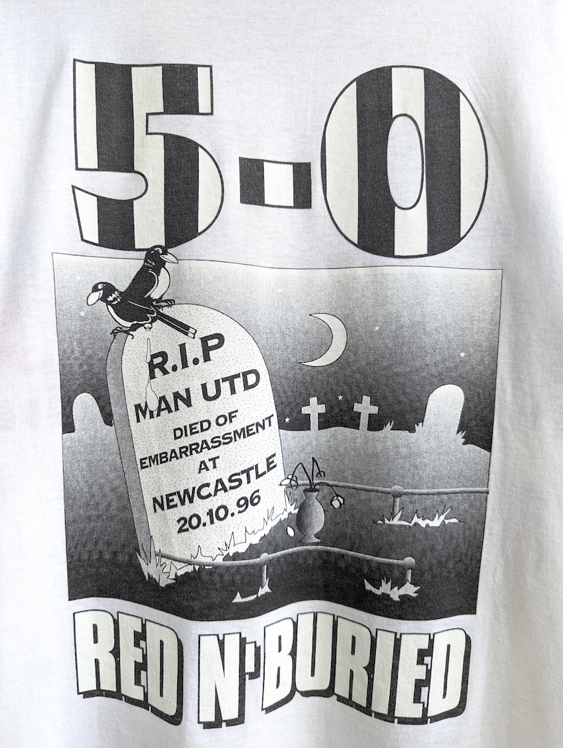 Pu&ograve; includere: Una maglietta bianca con una stampa grafica in bianco e nero. La grafica raffigura una lapide con il testo "R.I.P. Man Utd Died of Embarrassment at Newcastle 20.10.96" e due uccelli appollaiati in cima. Il testo "5-0 Red N' Buried" &egrave; stampato sotto la grafica.