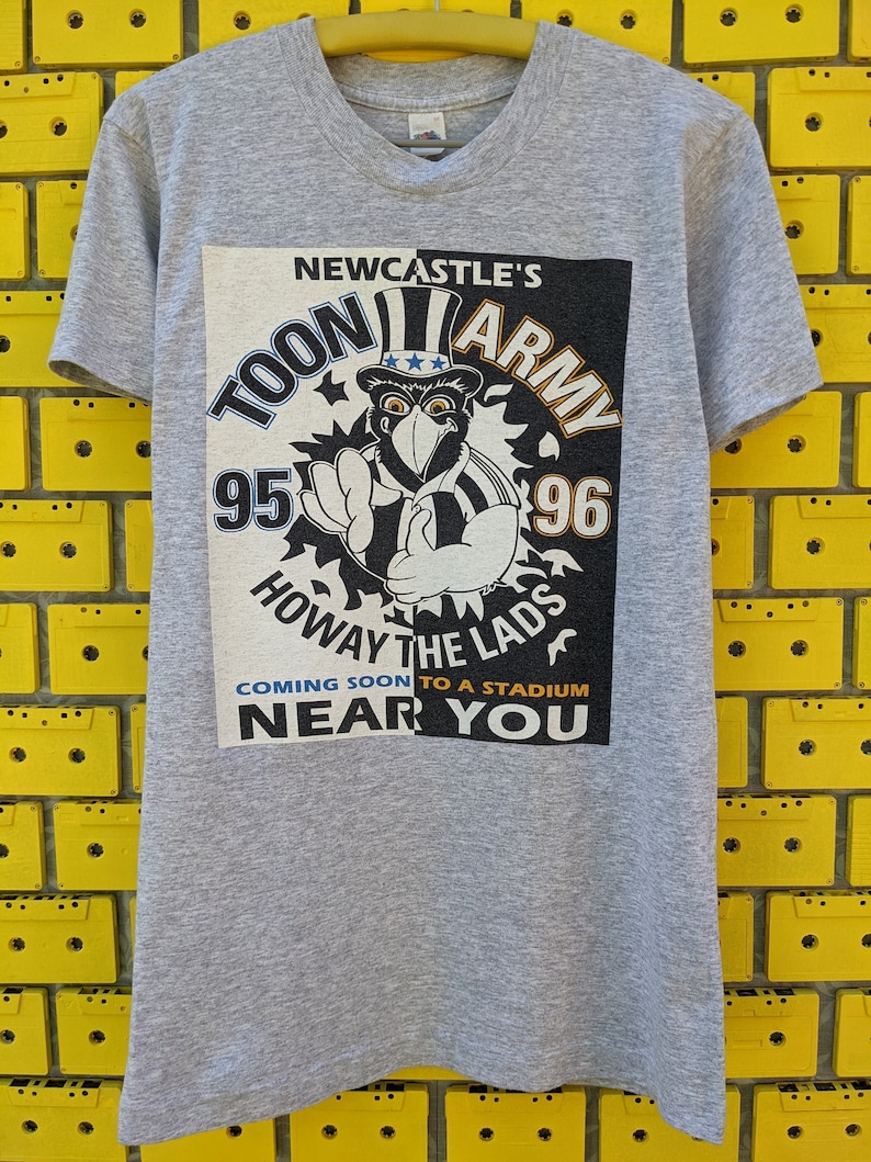 Puede incluir: Camiseta gris con un estampado gr&aacute;fico en blanco y negro de un p&aacute;jaro de dibujos animados con un sombrero con estrellas y rayas. El gr&aacute;fico dice "Newcastle's Toon Army 95-96 Howay the Lads Coming Soon to a Stadium Near You."