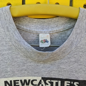 Puede incluir: Camiseta gris con un gr&aacute;fico en blanco y negro que dice "NEWCASTLE'S" en la parte delantera. La camiseta tiene una etiqueta de Fruit of the Loom con la talla "S4" en ella.
