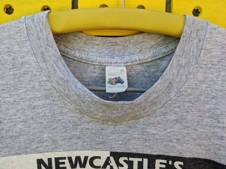 Puede incluir: Camiseta gris con un gr&aacute;fico en blanco y negro que dice "NEWCASTLE'S" en la parte delantera. La camiseta tiene una etiqueta de Fruit of the Loom con la talla "S4" en ella.