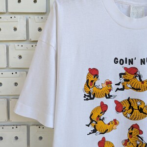 May include: White t-shirt with a graphic of cartoon peanuts in various poses. The text "GOIN' NUTS" is printed on the shirt.