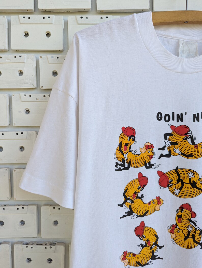 May include: White t-shirt with a graphic of cartoon peanuts in various poses. The text "GOIN' NUTS" is printed on the shirt.
