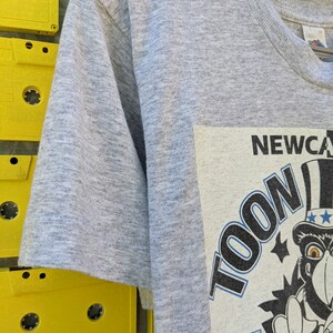 Puede incluir: Camiseta gris de manga corta con un estampado gr&aacute;fico de un personaje de dibujos animados con un sombrero de copa y sosteniendo una bandera. El texto "TOON 95" y "NEWCASTLE" son visibles en el gr&aacute;fico.