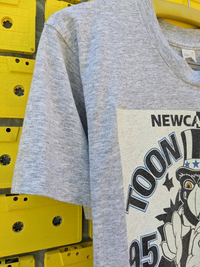 Puede incluir: Camiseta gris de manga corta con un estampado gr&aacute;fico de un personaje de dibujos animados con un sombrero de copa y sosteniendo una bandera. El texto "TOON 95" y "NEWCASTLE" son visibles en el gr&aacute;fico.