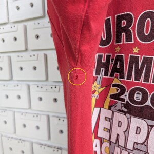 May include: Red t-shirt with a graphic design featuring the words "EURO HAM 2005" and "LIVERPOOL AC MILAN". The shirt has a crew neck and short sleeves. The background is a white wall with cassette tapes.