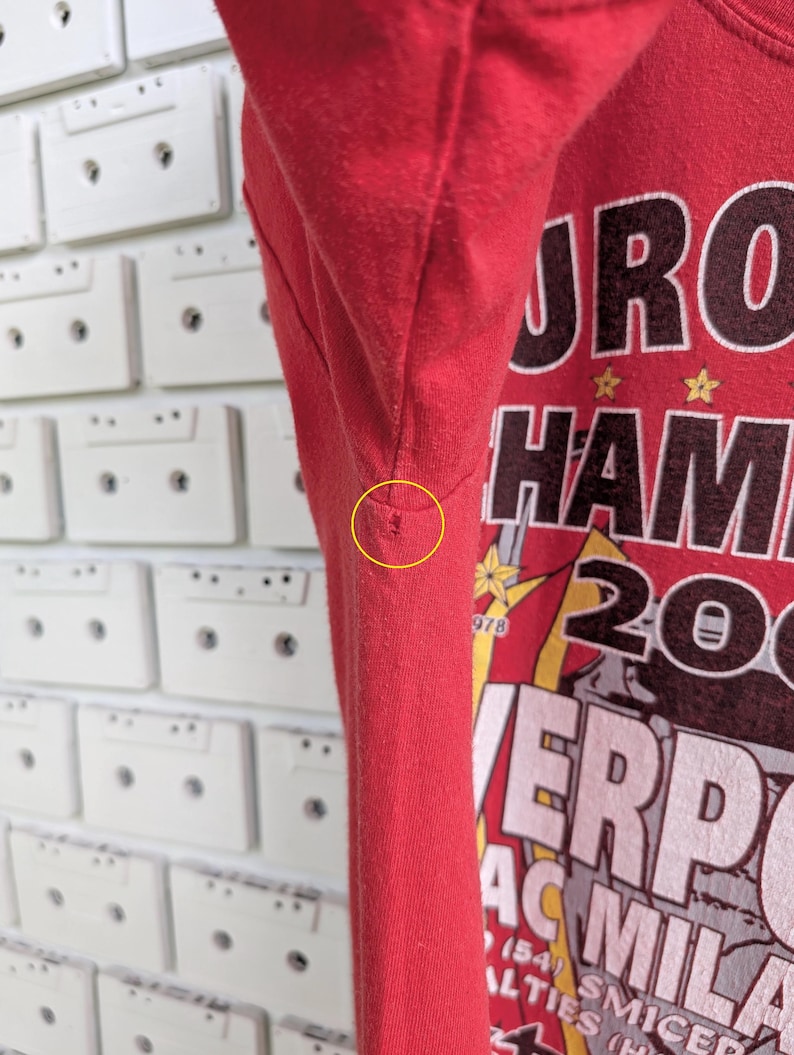 May include: Red t-shirt with a graphic design featuring the words "EURO HAM 2005" and "LIVERPOOL AC MILAN". The shirt has a crew neck and short sleeves. The background is a white wall with cassette tapes.
