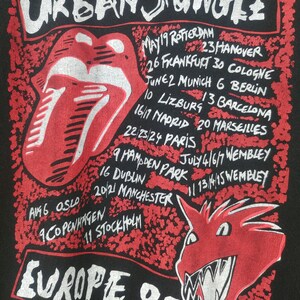 May include: Black t-shirt with a red and white graphic of a tongue and a dragon. The text "URBAN JUNGLE" is at the top of the graphic and "EUROPE 90" is at the bottom. The graphic also includes a list of cities and dates for a tour.