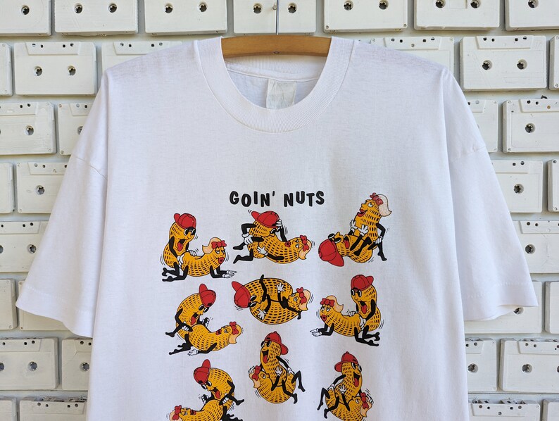 May include: White t-shirt with a graphic of peanuts in various poses with the text "Goin' Nuts" printed on the front.