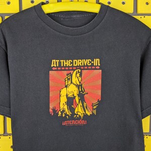 May include: Black t-shirt featuring a yellow and red graphic of a horse pulling a chariot. The text "At The Drive-In" and "&iexcl;Atenci&oacute;n!" is printed on the shirt.