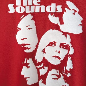 May include: Red t-shirt with a white graphic of the band "The Sounds" and four white silhouettes of people's faces.