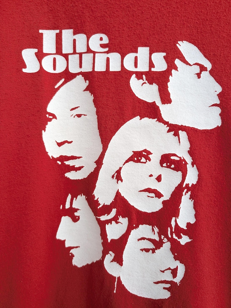 May include: Red t-shirt with a white graphic of the band "The Sounds" and four white silhouettes of people's faces.