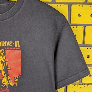 May include: Black t-shirt featuring a graphic of a woman holding a sword in front of a red and yellow sunburst. The text "Drive-In" is above the graphic and "enci&oacute;n" is below.