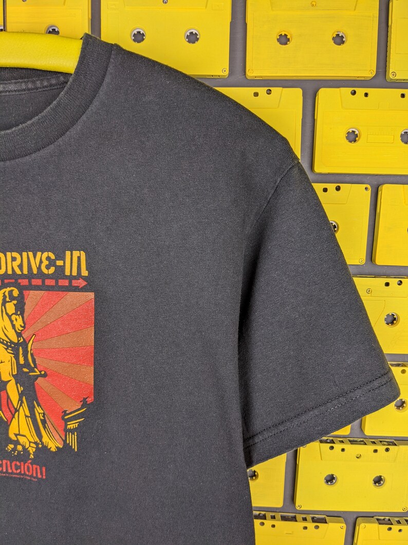 May include: Black t-shirt featuring a graphic of a woman holding a sword in front of a red and yellow sunburst. The text "Drive-In" is above the graphic and "enci&oacute;n" is below.