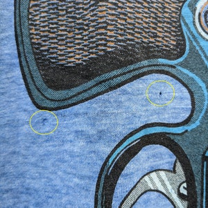 Puede incluir: Una camiseta azul con un estampado gr&aacute;fico negro y azul de un personaje de dibujos animados. El personaje tiene una cabeza grande y un cuerpo peque&ntilde;o. El personaje lleva un sombrero y tiene una nariz grande. La camiseta tiene dos peque&ntilde;os agujeros en la tela.
